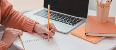 A person writing in a notebook beside a laptop