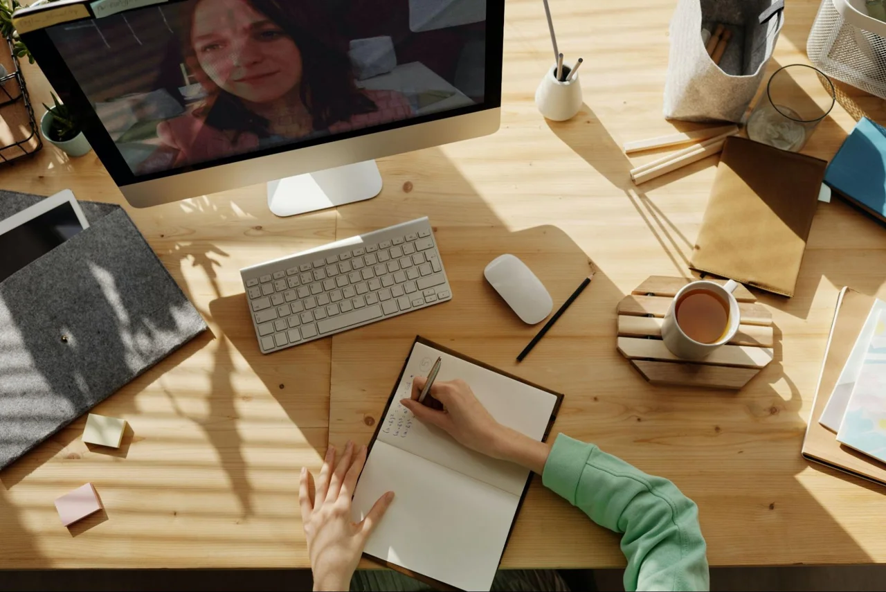 A person writing notes at a desk with a computer